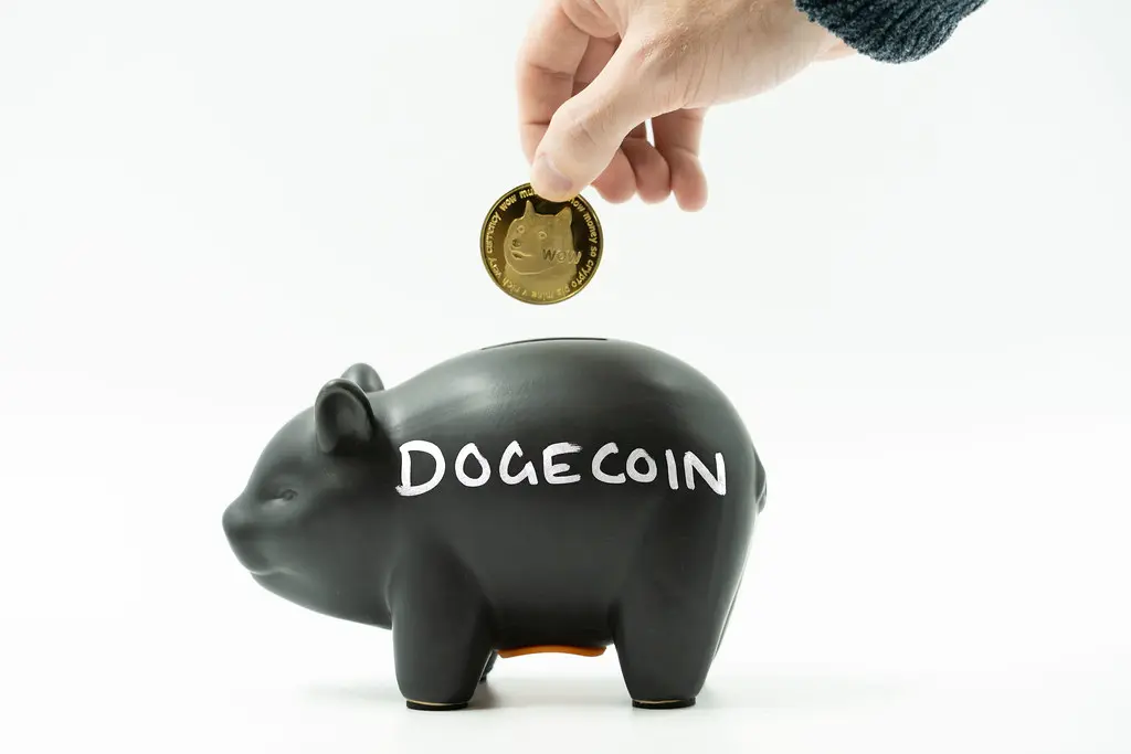 Uma mão coloca uma moeda dourada do Dogecoin em um cofrinho de porquinho preto com a palavra DOGECOIN escrita.