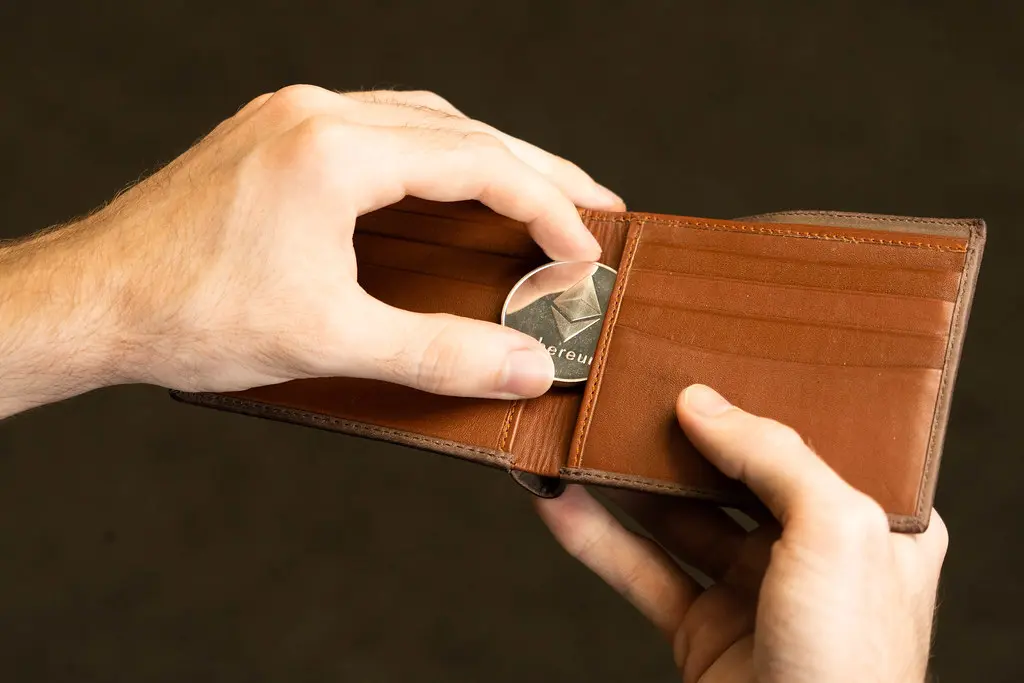 Mãos colocando uma moeda física do Ethereum em uma carteira de couro marrom.