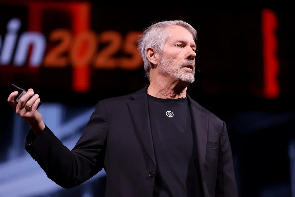 Michael Saylor falando em um palco, vestindo blazer preto e camiseta com o símbolo do Bitcoin.